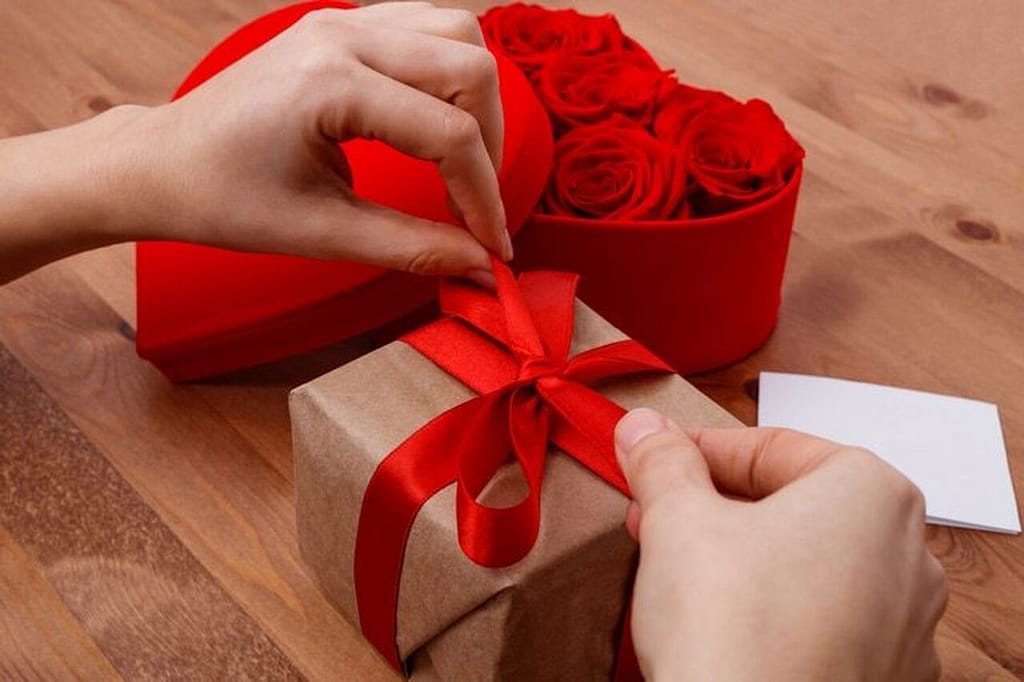 Pessoa abrindo ou fechando o laço de uma caixinha de presente, que tem uma caixa vermelha em formado de coração ao lado, junto a um bilhetinho em branco. A imagem simboliza ideias de brindes para dia dos namorados