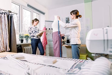 Duas mulheres produzindo roupas, uma desenhando e outra medindo uma manequim. No cômodo há também máquina de costura, desenhos de roupas na parede, uma janela com dois vasos de flores e um cabide (arara) com roupas, simbolizando mimos para clientes de roupas