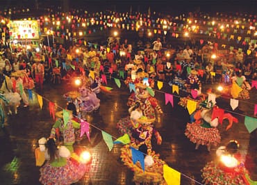 Pessoas com vestimentas típicas de festa junina celebrando em ambiente com bandeirinhas coloridas penduradas