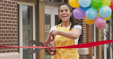 Balões de festa ao fundo. Mulher com tesoura cortando fita vermelha de inauguração, simbolizando o que dar de brinde em inauguração de loja