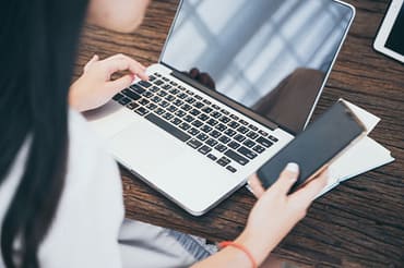 Mulher usando celular e notebook, simbolizando estratégia de branding. Há também um bloco de notas ao lado do notebook, também sobre a mesa de escritório.