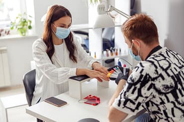 Simbolizando a dúvida de o que dar de mimos para clientes de manicure: Homem manicure de luvas analisando cores para unha de uma cliente, ambos estão de máscara num salão. A mesa de atendimento dele ter uma luminária e um apoio acolchoado para as mãos da cliente. Há um celular ao lado desse apoio.