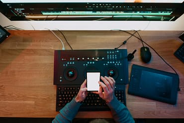 Pessoa mexendo em celular, com teclado multimídia, mesa digitalizadora e monitor de computador sobre a mesa.