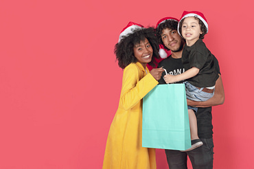 Uma família (mulher, e homem segurando um menino pequeno), todos com gorro de papai Noel. A mãe e a criança estão segurando uma grande sacola, que representa brinde para fidelizar clientes