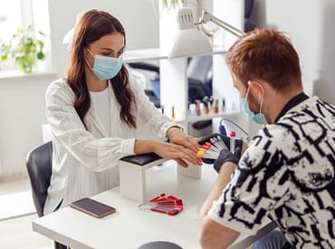 Simbolizando a dúvida de o que dar de mimos para clientes de manicure: Homem manicure de luvas analisando cores para unha de uma cliente, ambos estão de máscara num salão. A mesa de atendimento dele ter uma luminária e um apoio acolchoado para as mãos da cliente. Há um celular ao lado desse apoio.