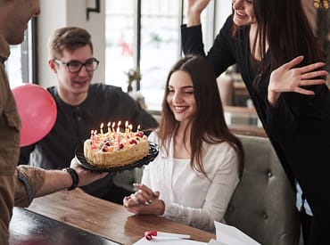 Funcionária sorrindo recebendo bolo de aniversário e colegas ao redor celebrando.