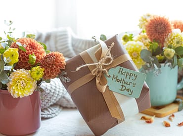 Simbolizando brinde dia das mães corporativo: Embrulho de caixa de presente com etiqueta escrito "Happy Mothers Day" (Feliz dia das mães, em inglês), com vasos de flores ao redor