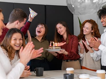 Simbolizando presentes para funcionários, um grupo de funcionários comemorando um aniversário com bolo, chapeuzinho de festa, balão e copos sobre a mesa