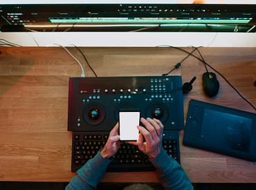 Pessoa mexendo em celular, com teclado multimídia, mesa digitalizadora e monitor de computador sobre a mesa.