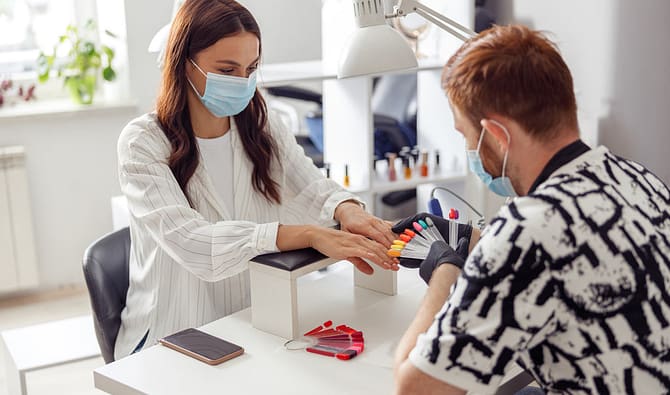 Simbolizando a dúvida de o que dar de mimos para clientes de manicure: Homem manicure de luvas analisando cores para unha de uma cliente, ambos estão de máscara num salão. A mesa de atendimento dele ter uma luminária e um apoio acolchoado para as mãos da cliente. Há um celular ao lado desse apoio.