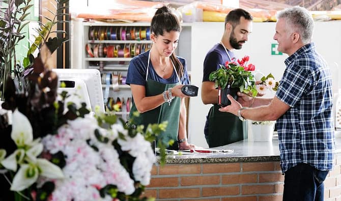 Operadora de caixa em floricultura segurando leitor de código de barras para um cliente segurando um vaso de flores, que representa uma opção entre presentes para clientes VIPs