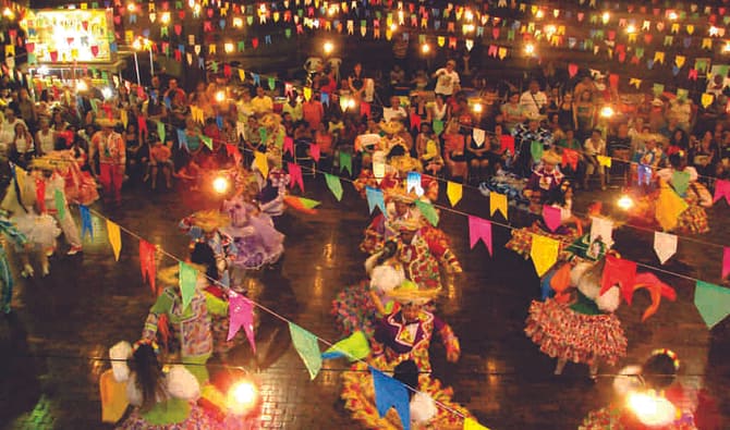 Pessoas com vestimentas típicas de festa junina celebrando em ambiente com bandeirinhas coloridas penduradas