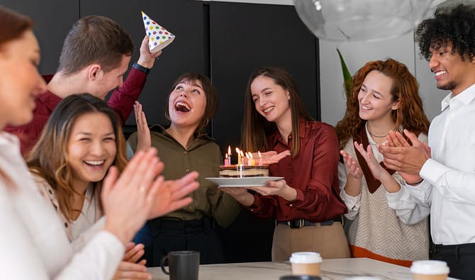 Simbolizando presentes para funcionários, um grupo de funcionários comemorando um aniversário com bolo, chapeuzinho de festa, balão e copos sobre a mesa