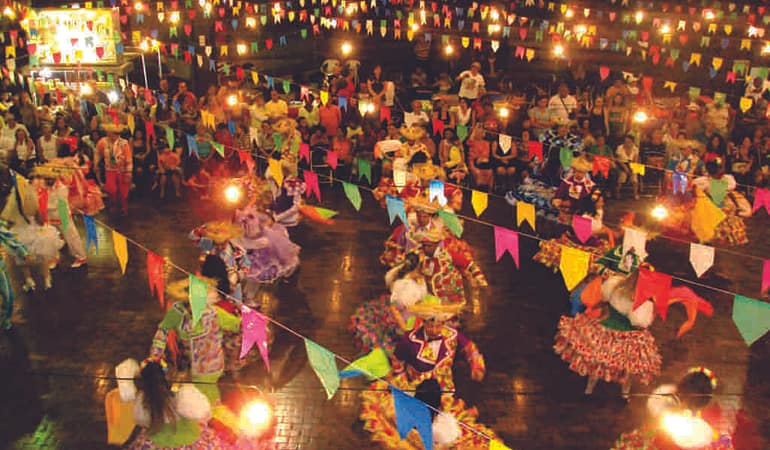 Pessoas com vestimentas típicas de festa junina celebrando em ambiente com bandeirinhas coloridas penduradas