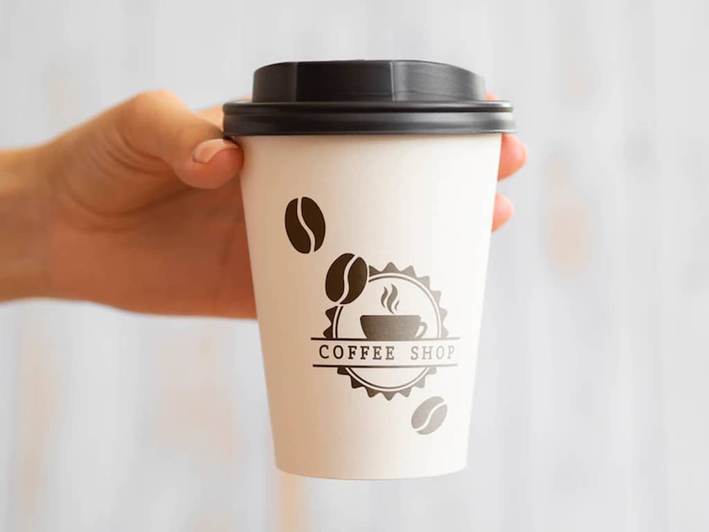 Mão segurando um copo com tampa personalizado com estampa de "Coffee shop" com logotipo e grãos de café, simbolizando a dúvida de como escolher o modelo ou tipo de copo personalizado ideal