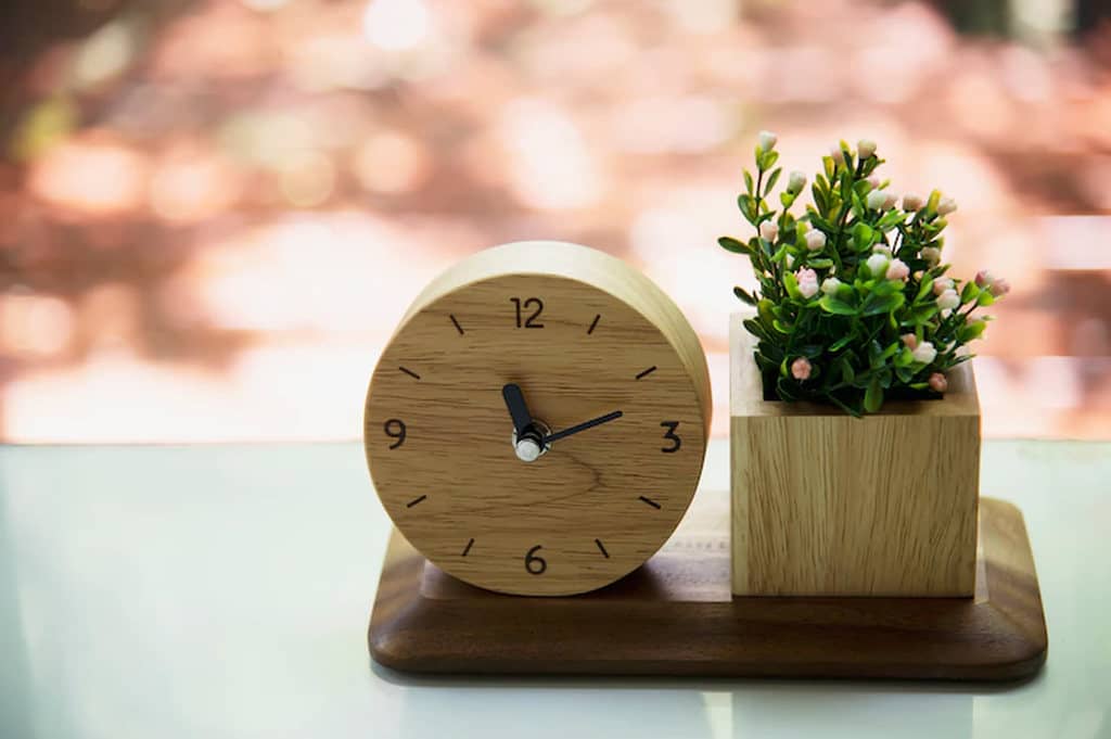 Relógio de madeira ao lado de vasinho de plantas de madeira, simbolizando o momento ideal de dar brindes para loja de roupas