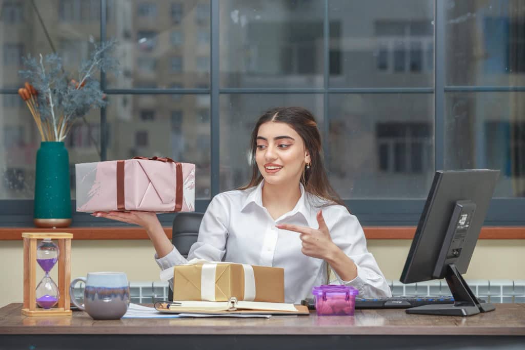 Mulher sorrindo, de camisa social branca, sentada em escritório segurando caixa de presente e apontando para a caixa com a outra mão, representando brindes uteis e criativos