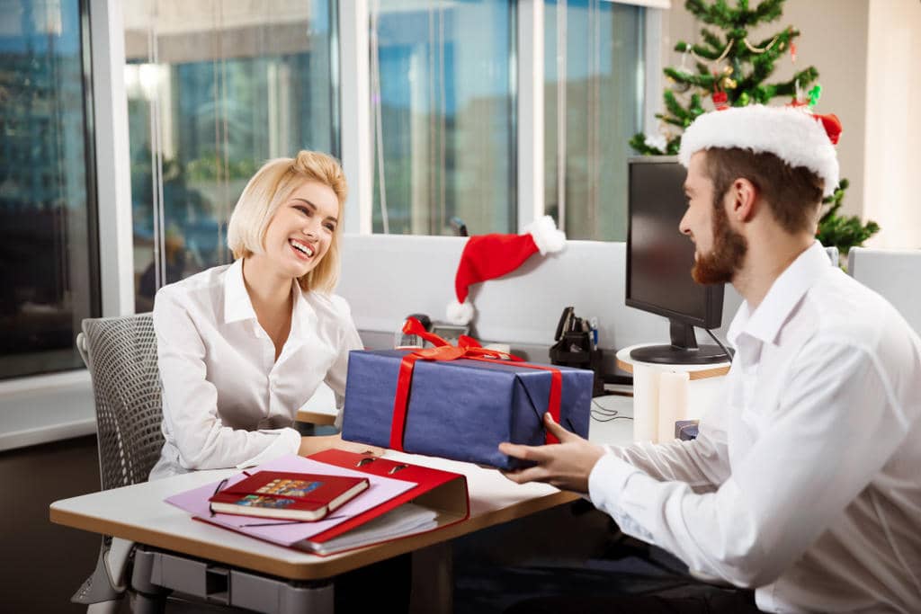 Um funcionário com touca de papai Noel entregando um presente para uma funcionária, estão no escritório dela que tem uma árvores de Natal de decoração, além de escrivaninha com notebook