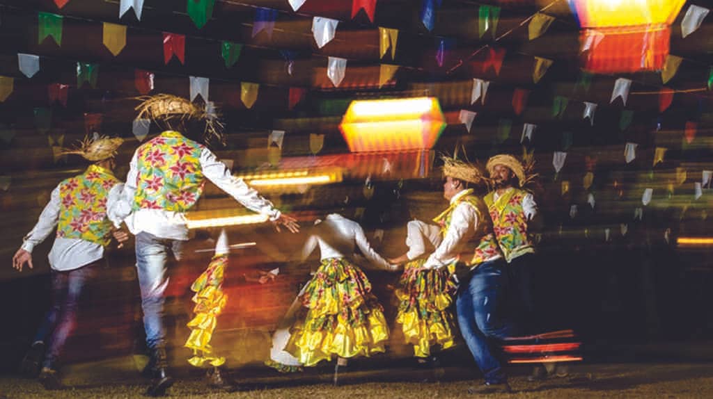 Pessoas com chaéu de palha dançando quadrilha e celebrando festa junina na empresa. Na decoração, há balões decorativos e bandeirinhas coloridas