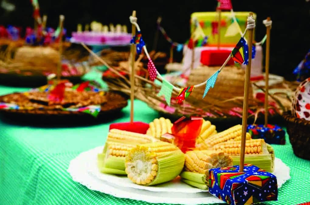 Mesa decorativa de festa junina na empresa com bandeirinhas coloridas e tecido xadrez. Milho e chapéus de palha sobre a mesa,