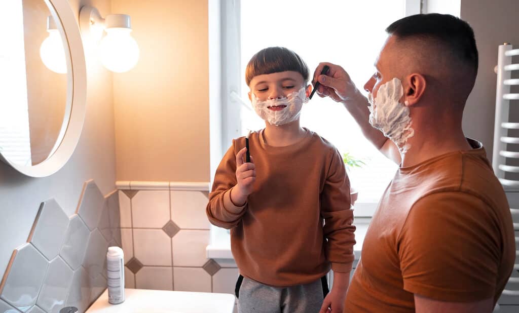 Filho e pai brincando de se barbear no banheiro. A imagem simboliza o brinde kit de barbear de dia dos pais corporativo