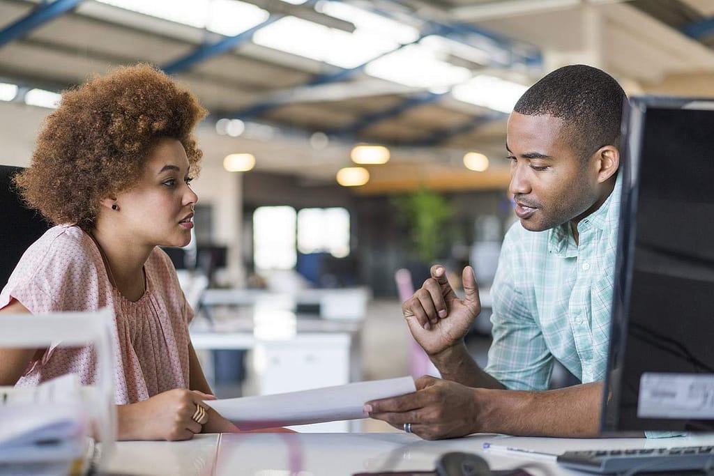 Homem e mulher interagindo, ele segurando uma folha de papel, demonstrando comunicação empresarial