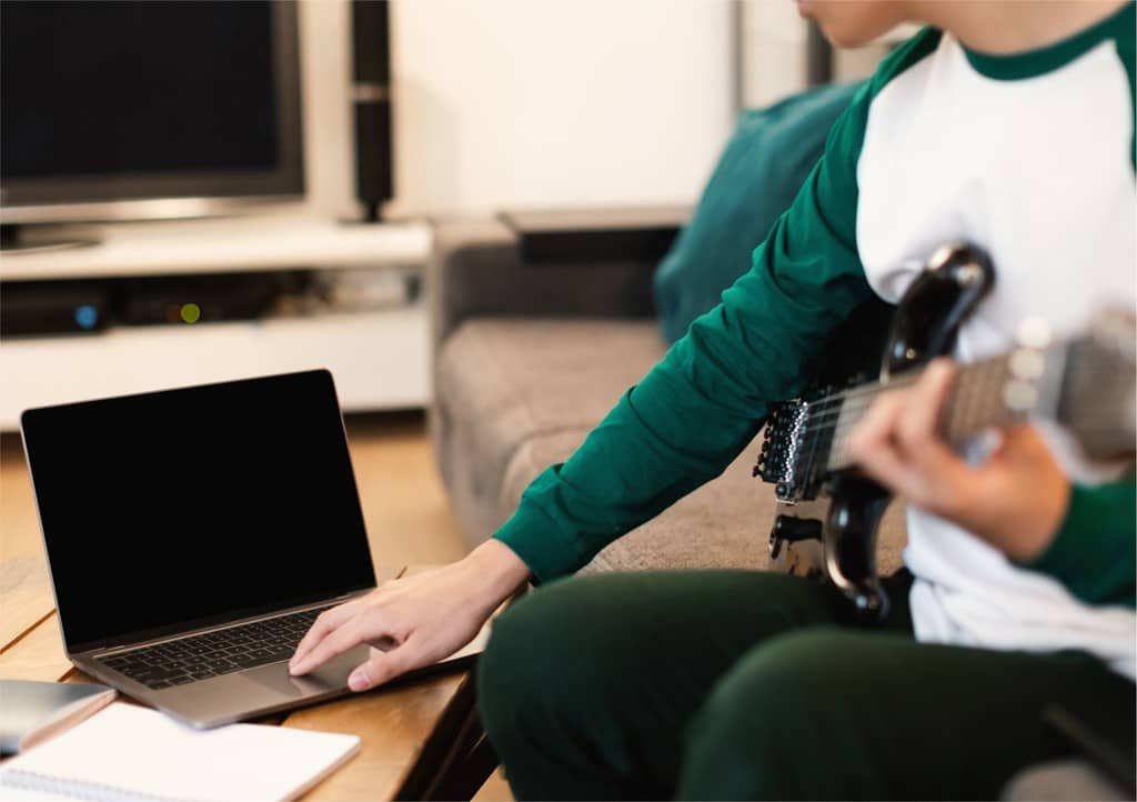 Jovem tocando guitarra sentado em sofá, enquanto mexe em notebook na mesa de centro, simbolizando a dúvida sobre o que dar de brinde para jovens conectados