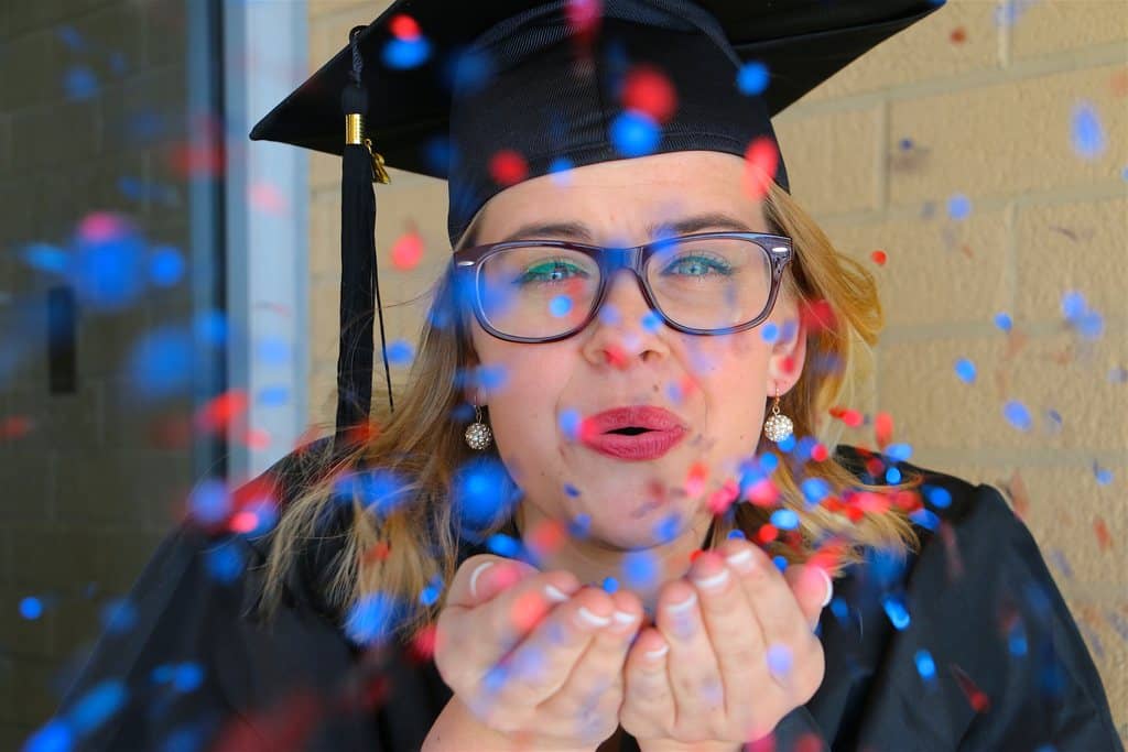 Mulher de óculos de grau, vestida com beca e capelo (chapéu de formatura), a mulher está feliz mandando beijo para câmera, e há confetes festivos no ar, simbolizando as melhores ideias de brindes para formatura