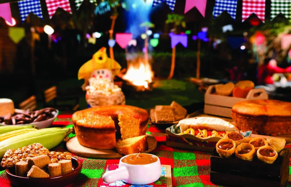 Mesa de festa junina na empresa com bolo, cones de doce de leite, panelinha com doce, paçoca, pé de moleque, doce de abóbora etc.