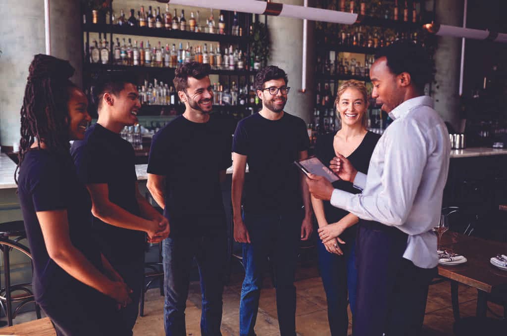 Lugar que vende bebidas com equipe motivada sorridente prestando atenção ao gestor que está falando e segurando um tablet