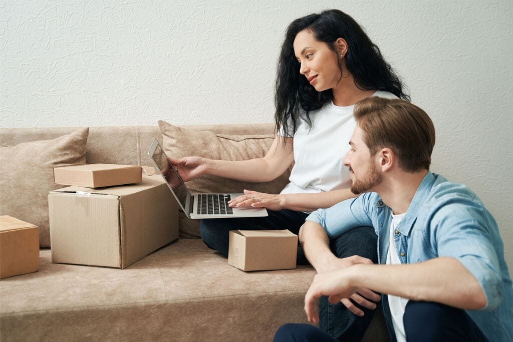 Casal de clientes comprando online, ela com o notebook no colo sentada no sofá, com caixas de papelão/embalagens ao redor, simbolizando brindes para o dia do cliente