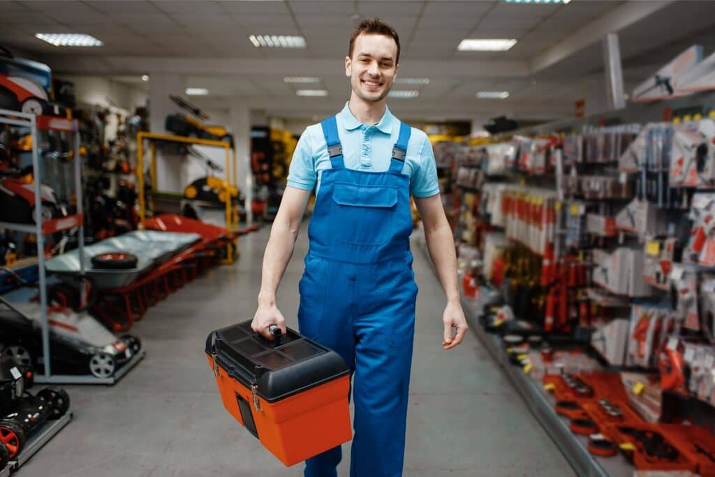 Vendedor de loja de ferramentas sorrindo largamente, caminhando no corredor segurando uma mala de ferramentas, que simboliza um kit de brindes para clientes