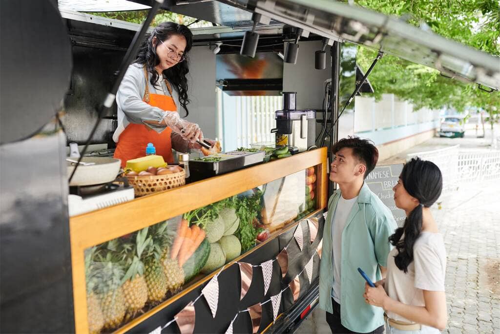 Mulher de avental atendendo em food truck que na vitrine tem cenouras, abacaxis e outros alimentos saudáveis. Dois clientes estão atentos ao que ela está preparando. a imagem simboliza mimos para fidelizar clientes